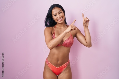 Hispanic woman wearing bikini smiling and looking at the camera pointing with two hands and fingers to the side.