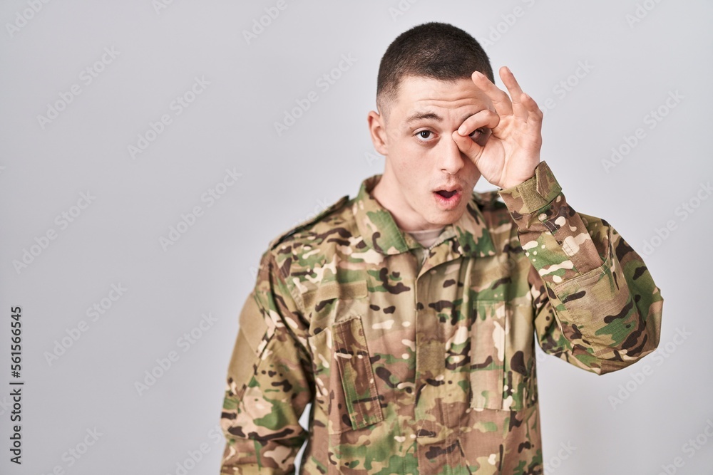 Young man wearing camouflage army uniform doing ok gesture shocked with surprised face, eye looking through fingers. unbelieving expression.