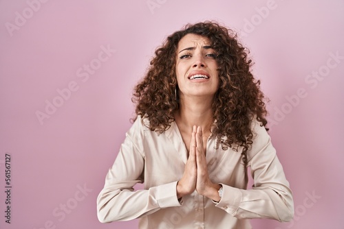 Fotografie Hispanic woman with curly hair standing over pink background begging and praying with hands together with hope expression on face very emotional and worried