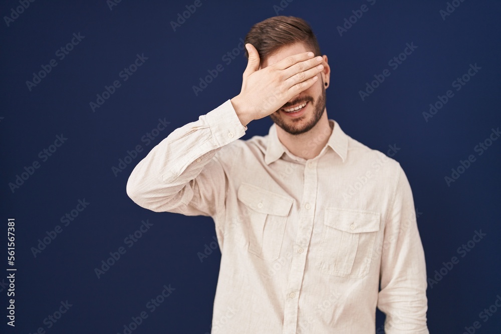 Hispanic man with beard standing over blue background smiling and laughing with hand on face covering eyes for surprise. blind concept. Hispanic man with beard standing over blue background smiling and laughing with hand on face covering eyes for surprise. blind concept.