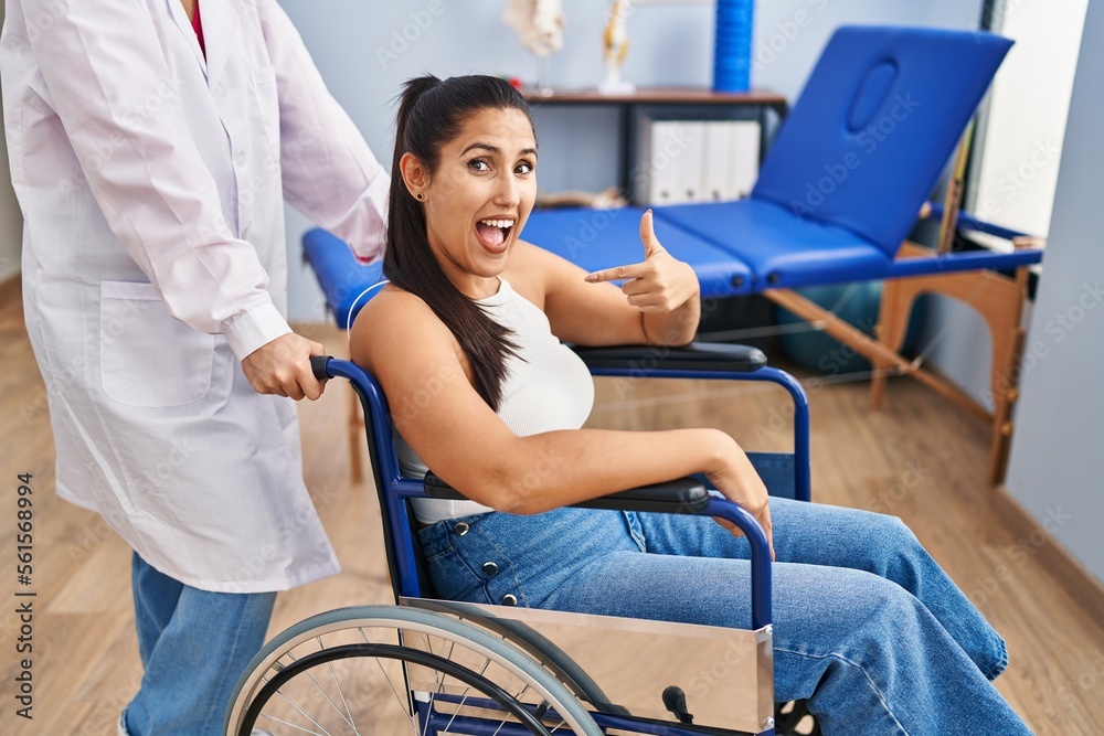Young hispanic woman sitting on wheelchair at physiotherapy clinic pointing finger to one self smiling happy and proud