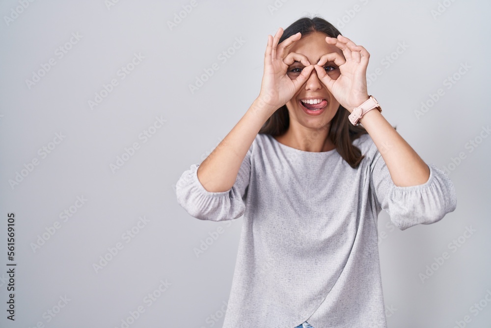 Young hispanic woman standing over white background doing ok gesture like binoculars sticking tongue out, eyes looking through fingers. crazy expression. Young hispanic woman standing over white background doing ok gesture like binoculars sticking tongue out, eyes looking through fingers. crazy expression.