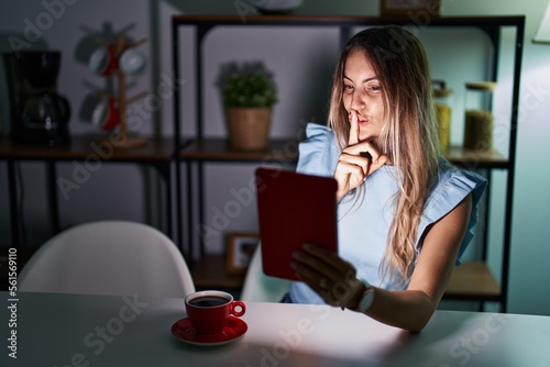 Young hispanic woman using touchpad sitting on the table at night asking to be quiet with finger on lips. silence and secret concept.