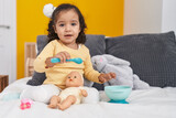 Adorable hispanic toddler playing with baby doll sitting on bed at bedroom
