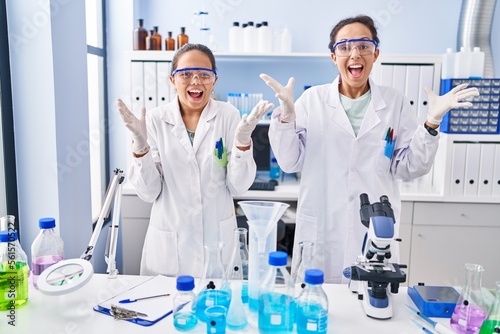 Young mother and daughter at scientist laboratory celebrating victory with happy smile and winner expression with raised hands
