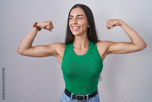 Young woman standing over isolated background showing arms muscles smiling proud. fitness concept.