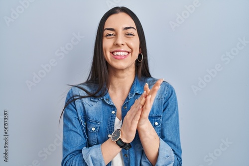 Hispanic woman standing over blue background clapping and applauding happy and joyful, smiling proud hands together