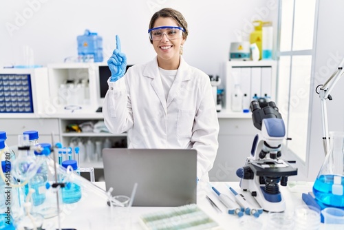 Young hispanic woman wearing scientist uniform working at laboratory surprised with an idea or question pointing finger with happy face  number one
