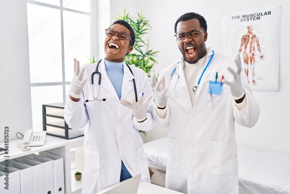 Young african american doctors working at medical clinic crazy and mad shouting and yelling with aggressive expression and arms raised. frustration concept.
