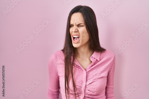 Young hispanic woman standing over pink background angry and mad screaming frustrated and furious  shouting with anger. rage and aggressive concept.