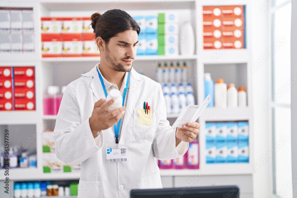 Young hispanic man pharmacist holding pills bottle reading prescription at pharmacy