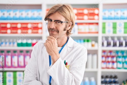 Caucasian man with mustache working at pharmacy drugstore looking confident at the camera smiling with crossed arms and hand raised on chin. thinking positive.