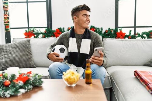 Young hispanic man supporting soccer game using smartphone sitting on sofa by christmas decor at home © Krakenimages.com