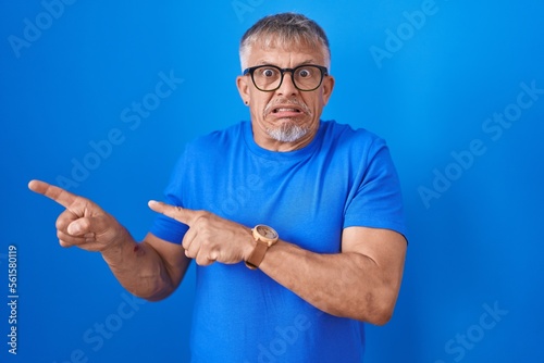 Hispanic man with grey hair standing over blue background pointing aside worried and nervous with both hands, concerned and surprised expression