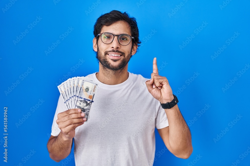 Handsome latin man holding dollars banknotes surprised with an idea or question pointing finger with happy face, number one