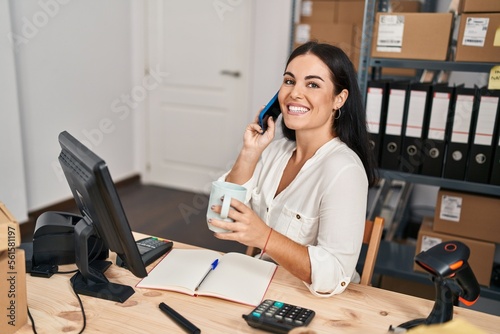 Young beautiful hispanic woman ecommerce business worker talking on smartphone drinking coffee at office