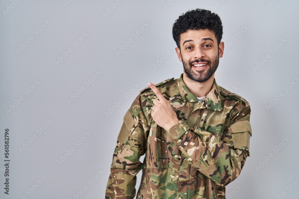 Arab man wearing camouflage army uniform with a big smile on face, pointing with hand finger to the side looking at the camera.