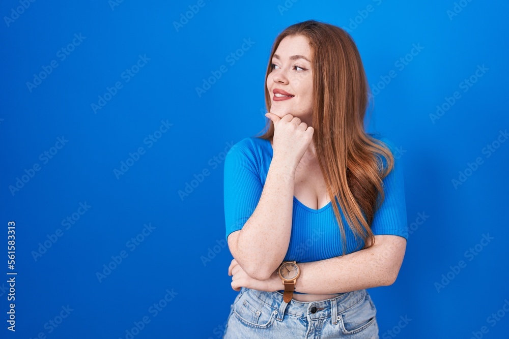 Redhead woman standing over blue background with hand on chin thinking about question, pensive expression. smiling and thoughtful face. doubt concept. Redhead woman standing over blue background with hand on chin thinking about question, pensive expression. smiling and thoughtful face. doubt concept.