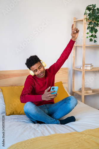 Cheerful black man using smartphone on bed listening music and dancing