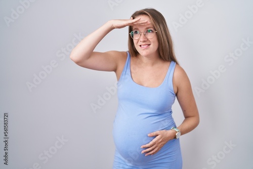 Young pregnant woman standing over white background very happy and smiling looking far away with hand over head. searching concept.