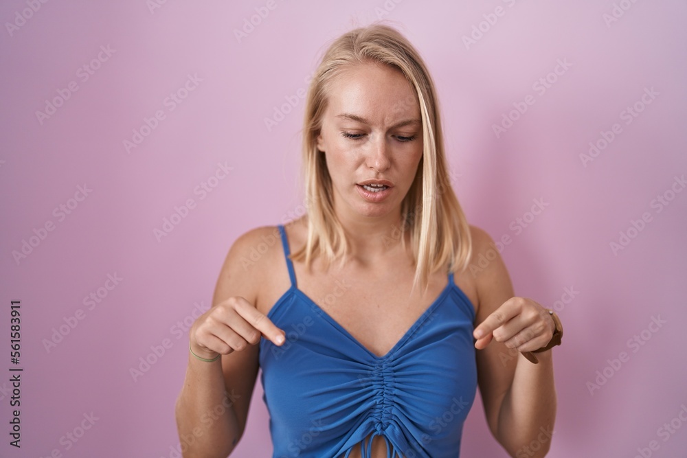 Young caucasian woman standing over pink background pointing down with fingers showing advertisement, surprised face and open mouth Young caucasian woman standing over pink background pointing down with fingers showing advertisement, surprised face and open mouth