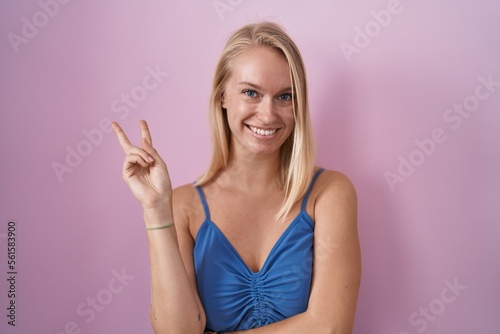 Young caucasian woman standing over pink background smiling with happy face winking at the camera doing victory sign with fingers. number two.