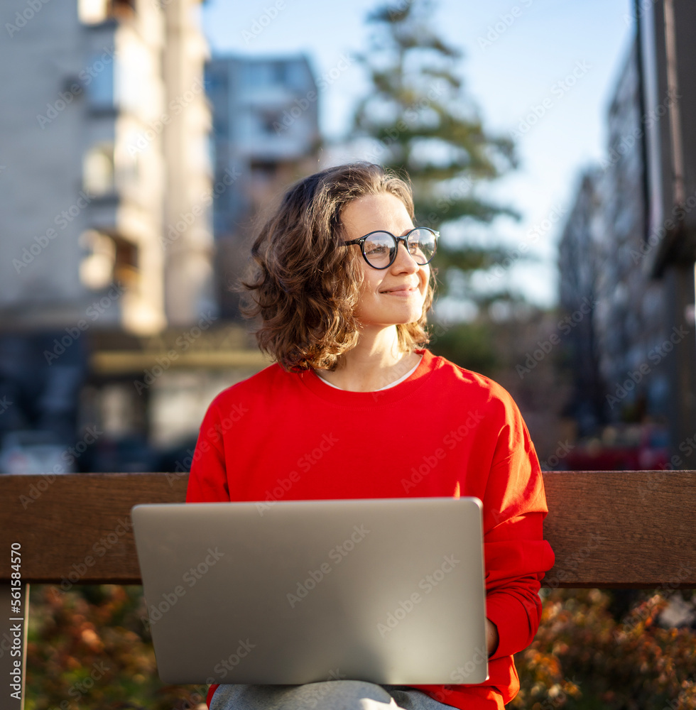 Young cheerful girl in glasses and a red blouse sitting on a bench in the city and using a laptop, online education and freelance concept