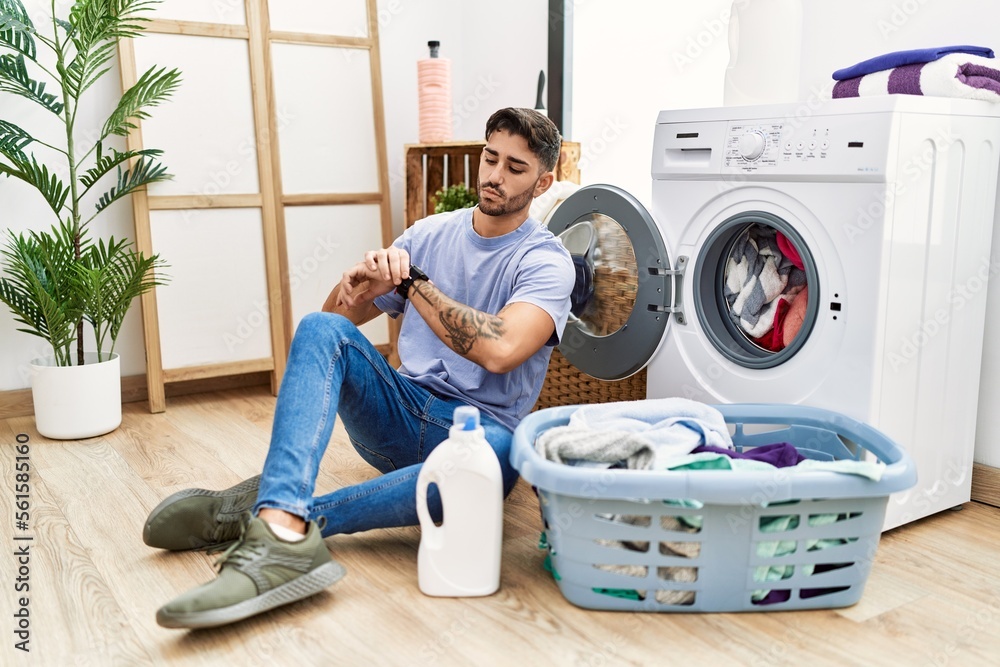 Young hispanic man putting dirty laundry into washing machine checking the time on wrist watch, relaxed and confident Young hispanic man putting dirty laundry into washing machine checking the time on wrist watch, relaxed and confident