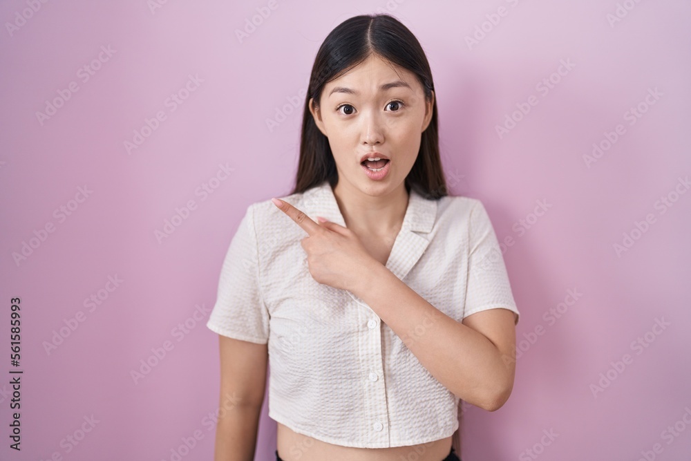 Chinese young woman standing over pink background surprised pointing with finger to the side, open mouth amazed expression. Chinese young woman standing over pink background surprised pointing with finger to the side, open mouth amazed expression.