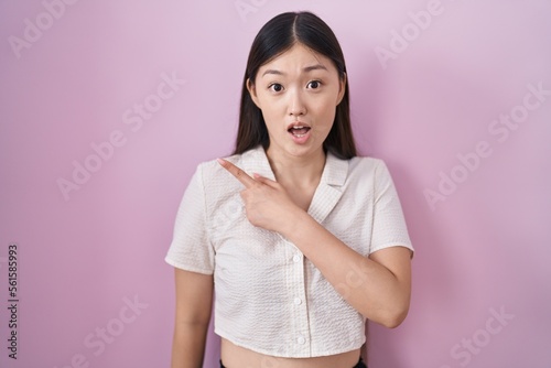 Chinese young woman standing over pink background surprised pointing with finger to the side, open mouth amazed expression.