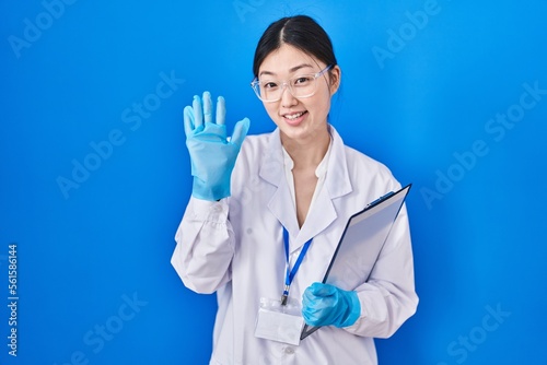 Chinese young woman working at scientist laboratory waiving saying hello happy and smiling  friendly welcome gesture