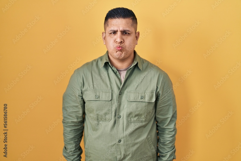 Fototapeta premium Hispanic young man standing over yellow background puffing cheeks with funny face. mouth inflated with air, crazy expression.