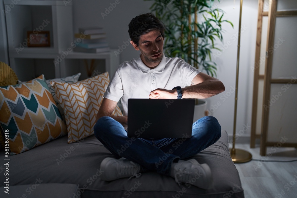 Young hispanic man using laptop at home at night checking the time on wrist watch, relaxed and confident Young hispanic man using laptop at home at night checking the time on wrist watch, relaxed and confident
