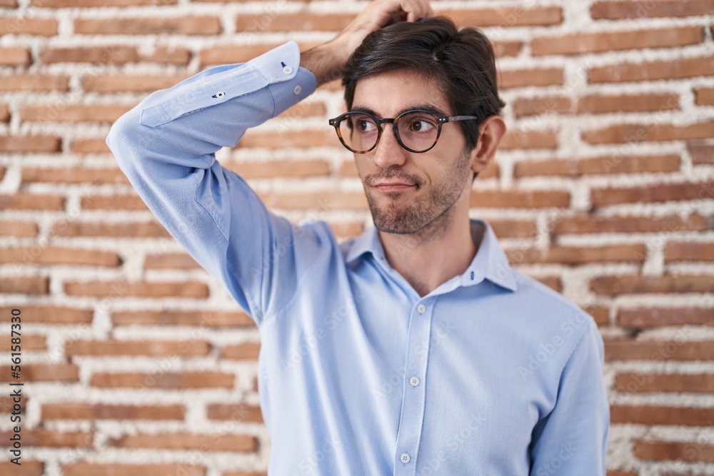 Young hispanic man standing over brick wall background confuse and wondering about question. uncertain with doubt, thinking with hand on head. pensive concept. Young hispanic man standing over brick wall background confuse and wondering about question. uncertain with doubt, thinking with hand on head. pensive concept.