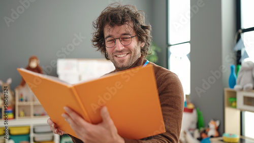 Young hispanic man preschool teacher reading book standing at kindergarten