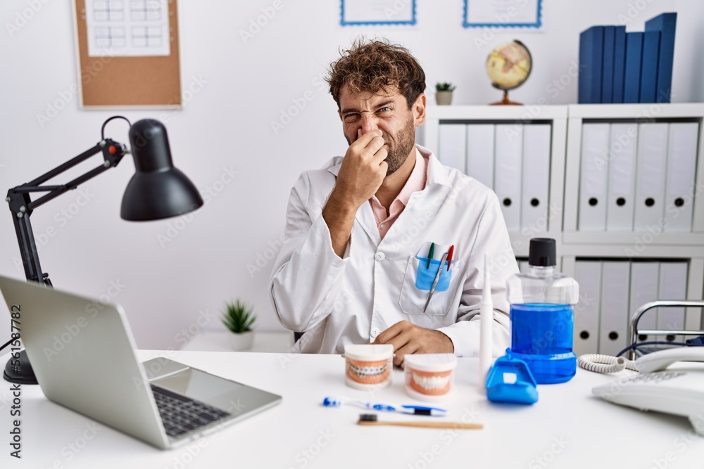 Young hispanic dentist man working at medical clinic smelling something