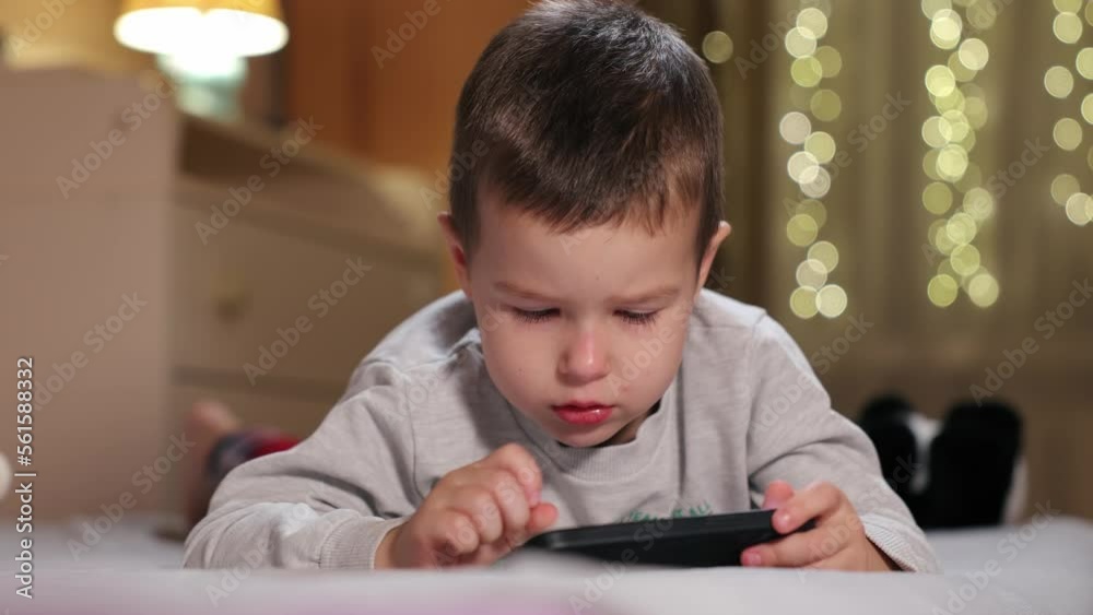 Cute Little Boy Uses Phone Lying On Bed. Kid Plays Mobile Games And Watch Video On Smartphone Cute Little Boy Uses Phone Lying On Bed. Kid Plays Mobile Games And Watch Video On Smartphone