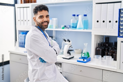 Young hispanic man wearing scientist uniform sitting with arms crossed gesture at laboratory