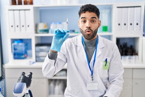Hispanic man with beard working at scientist laboratory holding syringe scared and amazed with open mouth for surprise  disbelief face