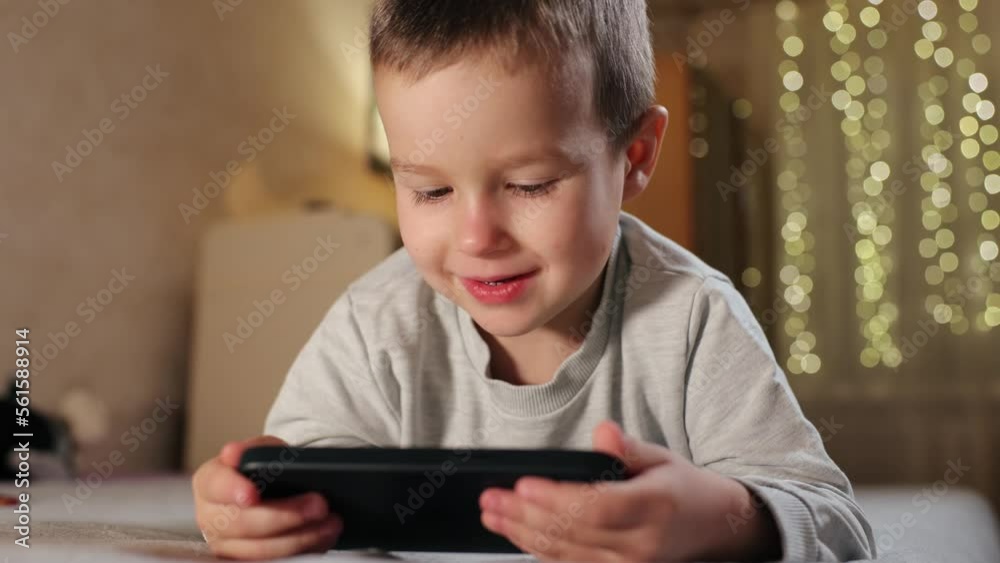 Cute Little Boy Uses Phone At Home in Room. Kid Plays Mobile Games On Smartphone Smiling and Showing Emotions Cute Little Boy Uses Phone At Home in Room. Kid Plays Mobile Games On Smartphone Smiling and Showing Emotions