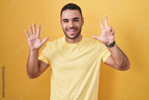 Young hispanic man standing over yellow background showing and pointing up with fingers number eight while smiling confident and happy. © Krakenimages.com
