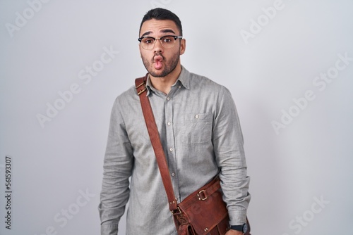 Young hispanic man wearing suitcase making fish face with lips, crazy and comical gesture. funny expression.