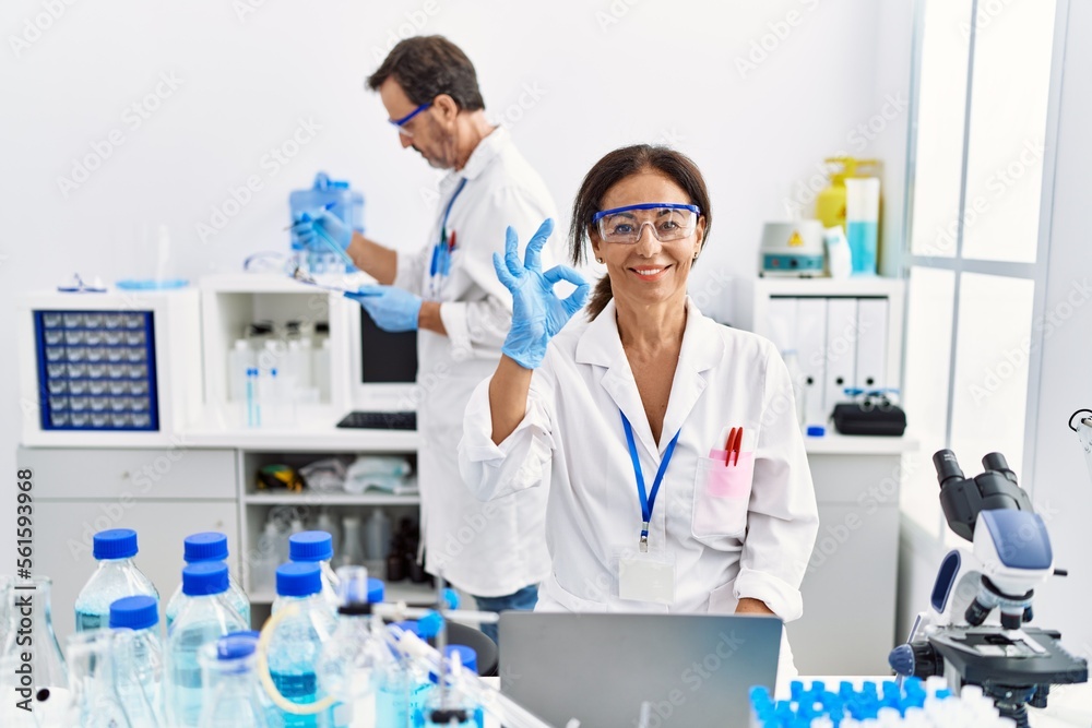 Middle age woman working at scientist laboratory doing ok sign with fingers, smiling friendly gesturing excellent symbol