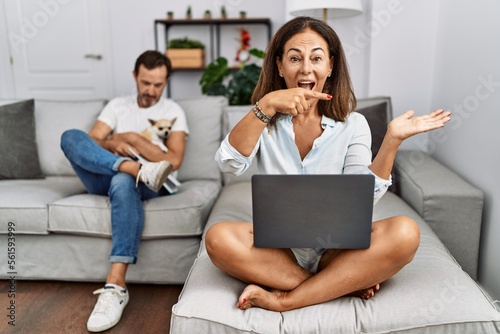 Hispanic middle age couple at home, woman using laptop amazed and smiling to the camera while presenting with hand and pointing with finger.