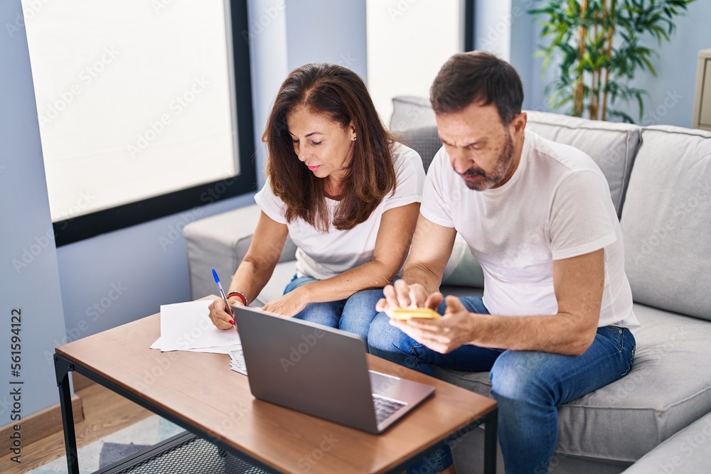 Middle age man and woman couple accounting using smartphone and laptop at home