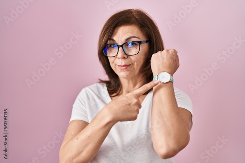 Middle age hispanic woman standing over pink background in hurry pointing to watch time  impatience  looking at the camera with relaxed expression