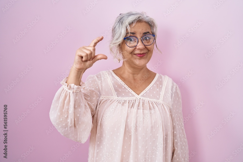 Middle age woman with grey hair standing over pink background smiling and confident gesturing with hand doing small size sign with fingers looking and the camera. measure concept.