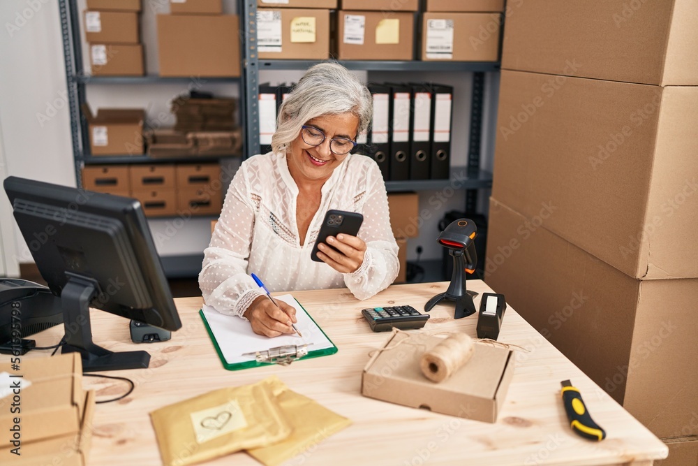Middle age woman ecommerce business worker using smartphone write on clipboard at office Middle age woman ecommerce business worker using smartphone write on clipboard at office
