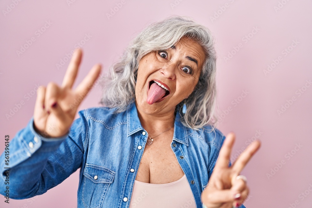 Middle age woman with grey hair standing over pink background smiling with tongue out showing fingers of both hands doing victory sign. number two. Middle age woman with grey hair standing over pink background smiling with tongue out showing fingers of both hands doing victory sign. number two.