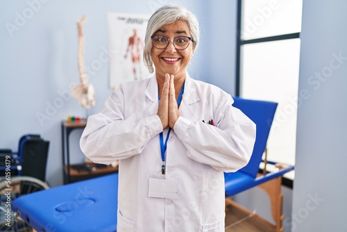 Middle age woman with grey hair working at pain recovery clinic praying with hands together asking for forgiveness smiling confident.